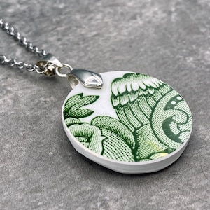 Puede incluir: Un collar de cadena de plata con un colgante redondo con un diseño floral verde y blanco.