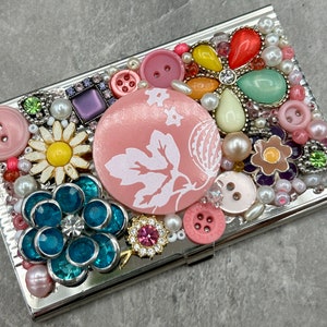 May include: A silver metal card holder decorated with colorful buttons, beads, and rhinestones. The design features a pink flower, a pink and white leaf, and a yellow flower.