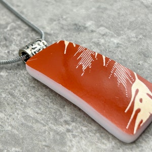 May include: A silver chain necklace with a rectangular pendant. The pendant is made of white and orange glass with a white abstract design.