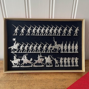 May include: A display case containing silver toy soldiers and cavalry figures. The figures are arranged on a black background and feature soldiers with rifles and riders on horseback. The case has a gold-colored frame.