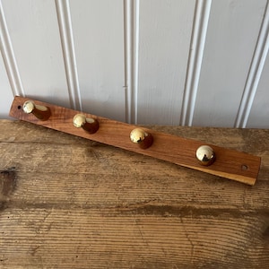 May include: A wooden coat rack with four brass ball hooks. The rack is made of a reddish-brown wood and is mounted on a white wall. The hooks are evenly spaced along the length of the rack. The rack is approximately 24 inches long.