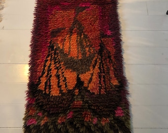 May include: A long, narrow rug with a shaggy texture. The rug features a colorful abstract design in shades of orange, pink, brown, and black. The design resembles a stylized bird or animal.