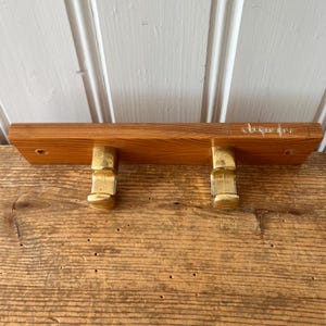 Puede incluir: Un perchero de pared de madera con dos ganchos de latón. La madera es de color marrón claro y los ganchos de latón son dorados. El perchero está montado sobre una superficie de madera.