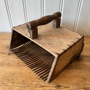 May include: A vintage wooden berry picker with a handle and metal tines. The wooden frame is rectangular with a curved front. The handle is attached to the top with two wooden supports. The text "Van Doren" is visible on the side.