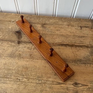May include: A wooden coat rack with five rounded pegs. The rack is made of a light brown wood and is rectangular in shape. The pegs are evenly spaced along the length of the rack. A simple and functional design.