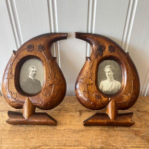 Pode incluir: Um par de molduras de madeira castanho-escura em forma de lágrima ou paisley. Cada moldura apresenta detalhes esculpidos de folhas e flores e uma abertura oval central contendo um retrato fotográfico a preto e branco de uma pessoa.