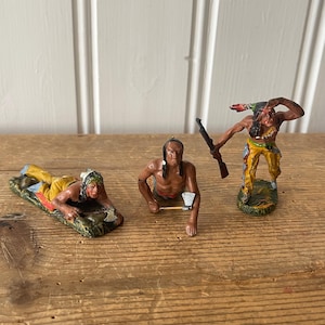 May include: Three painted metal figurines of Native American warriors. The figures are posed in different stances, one is holding a tomahawk, one is holding a rifle, and one is crawling on the ground. The figures are painted in shades of brown, yellow, and red.