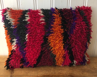 May include: A colorful, rectangular throw pillow with a shaggy, textured surface. The pillow features a geometric pattern in shades of red, orange, purple, black, and white.