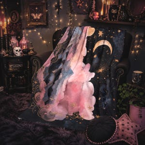 May include: A rectangular throw blanket featuring a dark navy blue night sky background with a large golden crescent moon and scattered stars. A prominent section of soft pink and light blue watercolor-style clouds flows across the center.