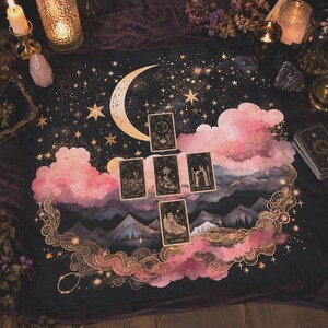 May include: A black tarot cloth with a celestial design featuring a crescent moon, stars, and pink clouds. Several tarot cards are laid out on the cloth. The design includes gold accents and a mountain range.
