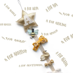 Puede incluir: Una estrella dorada con purpurina con la letra A en ella cuelga de una cinta blanca. La cinta está decorada con una abeja, un cisne y dos lazos. La estrella está rodeada de texto que dice "A para" seguido del nombre de una niña.