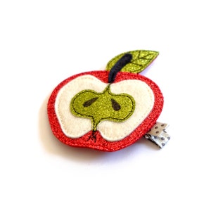 Könnte beinhalten: Ein roter Glitzerapfel mit einem grünen Filzkern und einem schwarzen Filzstiel. Der Apfel ist an einer silbernen Haarklammer mit einem schwarz-weißen Tupfenmuster befestigt.