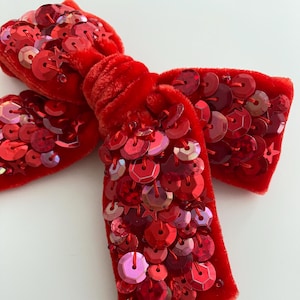 Könnte beinhalten: Eine rote Samtschleife mit roten Pailletten. Die Schleife ist zu einem Knoten gebunden und hat eine lange, fließende Schleife.