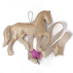 Könnte beinhalten: Ein goldener Glitzer-Pferde-Anhänger mit einem beigefarbenen Band und zwei sternförmigen Verzierungen, eine rosa und eine goldene.