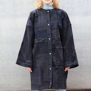 Può includere: Una lunga giacca di jeans nera con un design patchwork. La giacca ha una chiusura con bottoni sul davanti e grandi tasche.