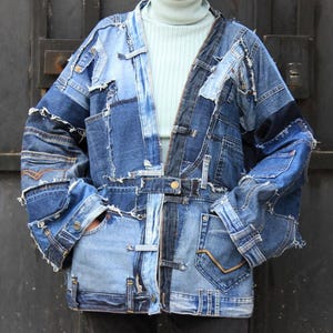 May include: A blue denim jacket made from patchwork denim with frayed edges. The jacket has a long, relaxed fit and a collarless design. The jacket is fastened with a button closure and has two pockets on the front.