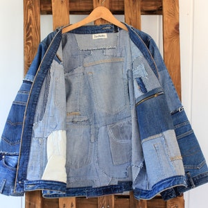 May include: A blue denim jacket with a patchwork design. The jacket is unzipped and the inside is lined with different shades of blue denim. The jacket has a zippered pocket on the right side.