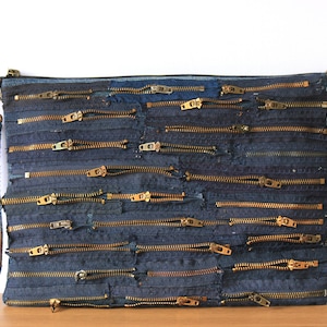 May include: A rectangular denim clutch bag featuring a horizontal patchwork design composed of multiple dark blue fabric strips. The surface is densely covered with numerous brass-colored metal zippers arranged in rows, complete with a matching wrist strap.