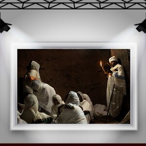 May include: A group of people wearing white robes gather in a dimly lit room. One person holds a book and a lit candle, while others sit on the floor. The room has a rustic, earthy feel.
