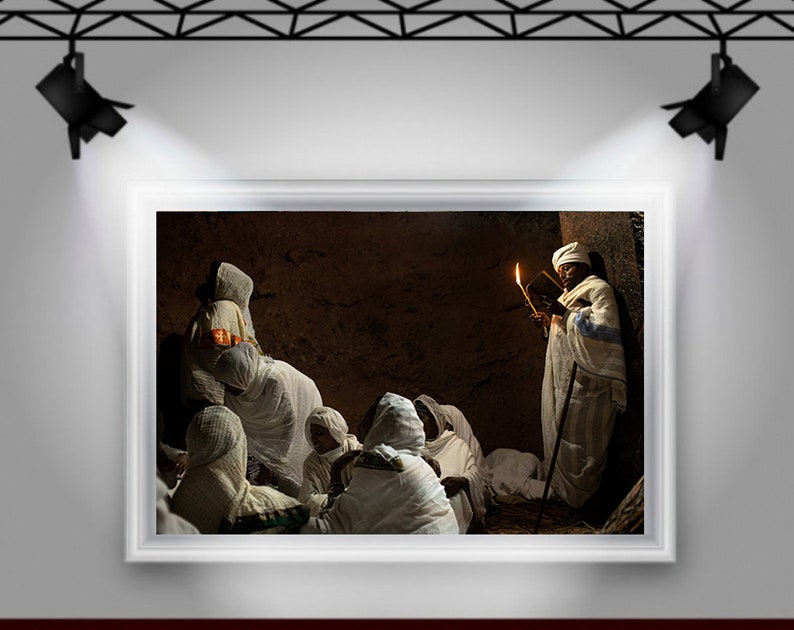 May include: A group of people wearing white robes gather in a dimly lit room. One person holds a book and a lit candle, while others sit on the floor. The room has a rustic, earthy feel.