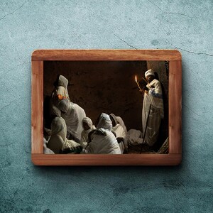 May include: A group of people wearing white robes gather in a dimly lit room. A person holding a book and a lit candle stands at the back of the group.