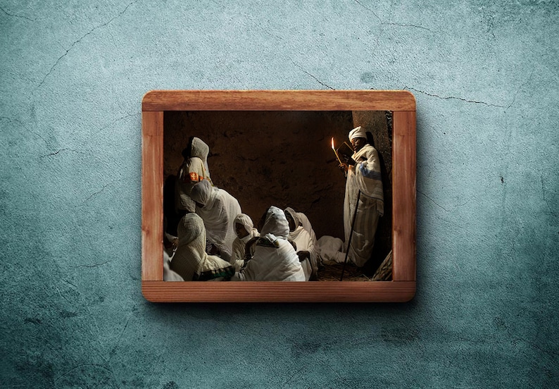 May include: A group of people wearing white robes gather in a dimly lit room. A person holding a book and a lit candle stands at the back of the group.