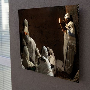 May include: A group of people wearing white robes gather in a dimly lit room. One person holds a lit candle and a book, while others sit and listen. The room has a rustic, earthy feel.