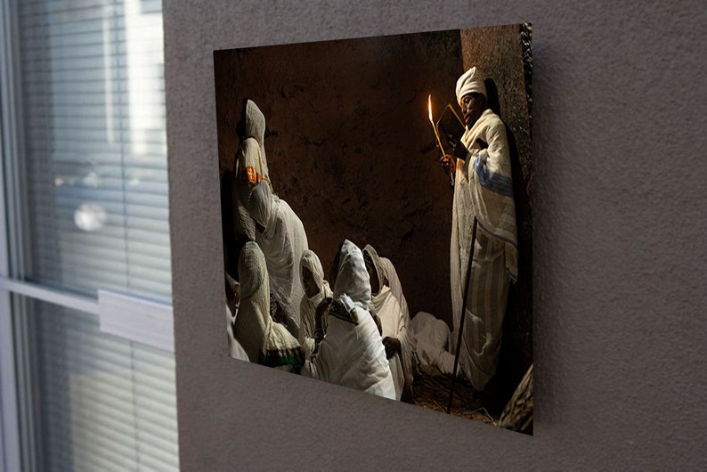 May include: A group of people wearing white robes gather in a dimly lit room. One person holds a lit candle and a book, while others sit and listen. The room has a rustic, earthy feel.