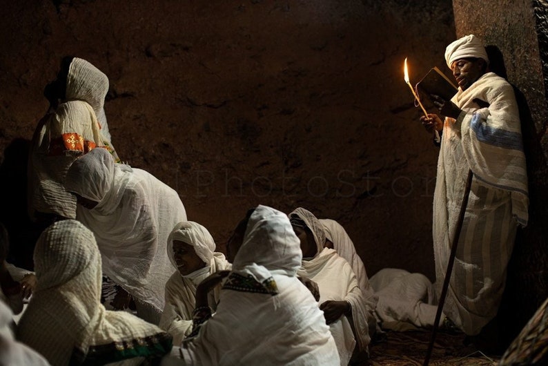 May include: A group of people wearing white robes gather in a dimly lit room. One person holds a lit candle and a book, while others sit on the floor. The room has a rough, stone wall in the background.