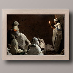 May include: A group of people wearing white robes gather in a dimly lit room. A person in the center holds a book and a lit candle, illuminating the scene.