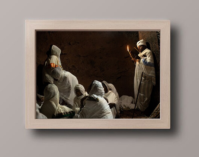 May include: A group of people wearing white robes gather in a dimly lit room. A person in the center holds a book and a lit candle, illuminating the scene.