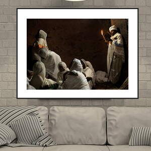 May include: A group of people wearing white robes gather in a dimly lit room. A person holding a lit candle stands in the back, reading from a book.