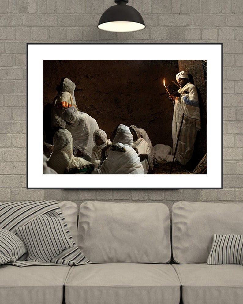 May include: A group of people wearing white robes gather in a dimly lit room. A person holding a lit candle stands in the back, reading from a book.