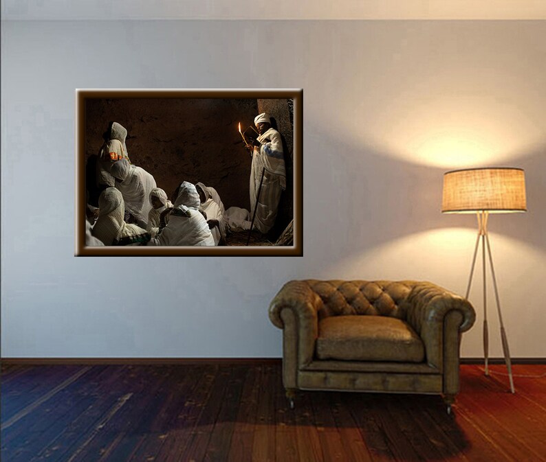 May include: A group of people wearing white robes gather in a dimly lit room. One person holds a lit candle and stands in the center of the group. The room has a stone wall and a wooden door.