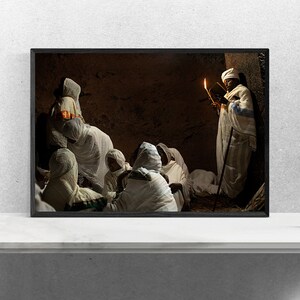 May include: A group of people wearing white robes gather in a dimly lit room. One person holds a lit candle and a book, while others sit on the floor listening. The room has a stone wall and a dark wooden door.