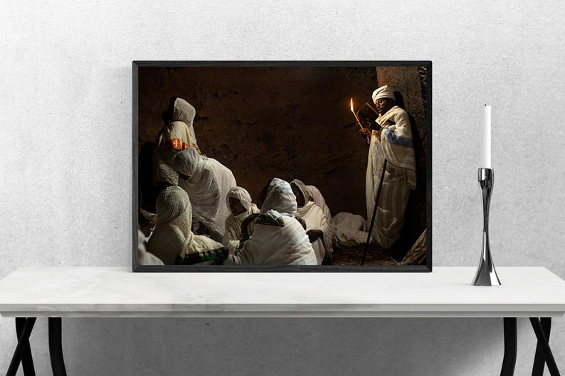 May include: A group of people wearing white robes gather in a dimly lit room. One person holds a lit candle and a book, while others sit on the floor listening. The room has a stone wall and a dark wooden door.
