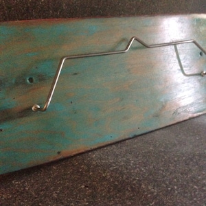 May include: A wall-mounted skateboard rack made of a distressed teal and brown wooden plank. A silver metal rack is attached to the wood. The rack is designed to hold a skateboard. The wood has a rustic, weathered appearance.