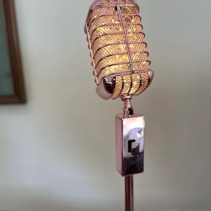 Könnte beinhalten: Eine Tischlampe in Form eines roségoldenen Mikrofons mit einem Drahtkäfig und einem Lichtschalter am Ständer.
