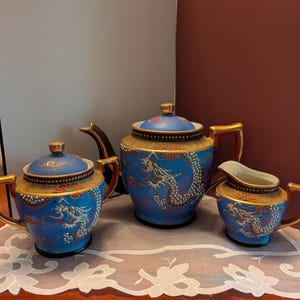 Peut inclure: Service à thé en porcelaine bleu et or comprenant une théière, un sucrier et un pot à crème. Chaque pièce est ornée d'un motif de dragon détaillé en blanc et rouge, avec des accents dorés sur les anses et les bords. L'ensemble est présenté sur un tissu en dentelle blanche.