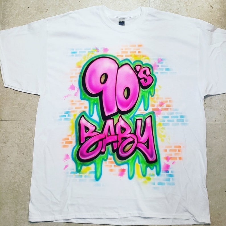 May include: White t-shirt with a graffiti design featuring the words "90's Baby" in pink and green with a brick wall background.