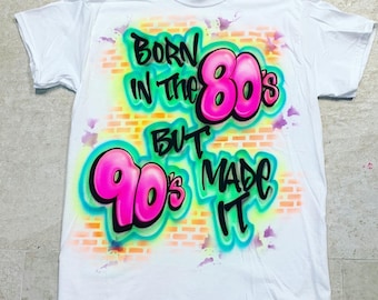 May include: White t-shirt with a graffiti-style design featuring the text "Born in the 80's but 90's made it". The design is in pink, green, and yellow on a brick wall background.