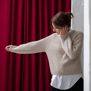 Könnte beinhalten: Eine Frau trägt einen beigefarbenen Strickpullover mit einem weißen Hemd darunter. Sie steht vor einem roten Vorhang und einer weißen Wand.