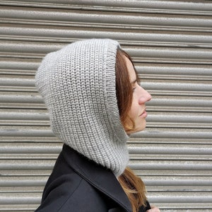 May include: A gray knitted hooded scarf with a ribbed texture. The scarf is worn by a person with long brown hair. The person is standing in front of a gray metal surface.