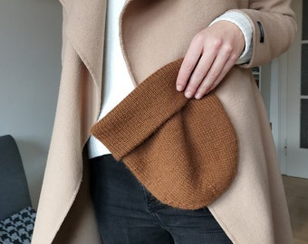 May include: A brown knit beanie hat being held by a person wearing a beige coat and black jeans.