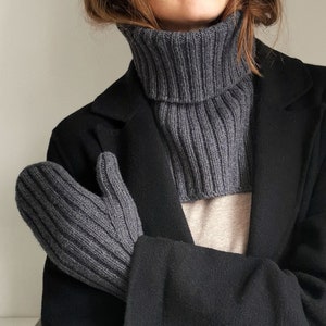 Könnte beinhalten: Ein grauer Rollkragenpullover mit gerippter Struktur. Der Pullover ist am Hals umgeklappt und wird unter einem schwarzen Mantel getragen.