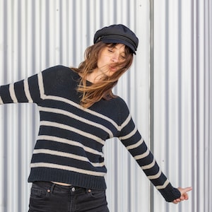 以下が含まれることがあります： 黒い帽子をかぶった、白黒のストライプのセーターを着た女性。女性は銀色の波板の壁の前に立っている。