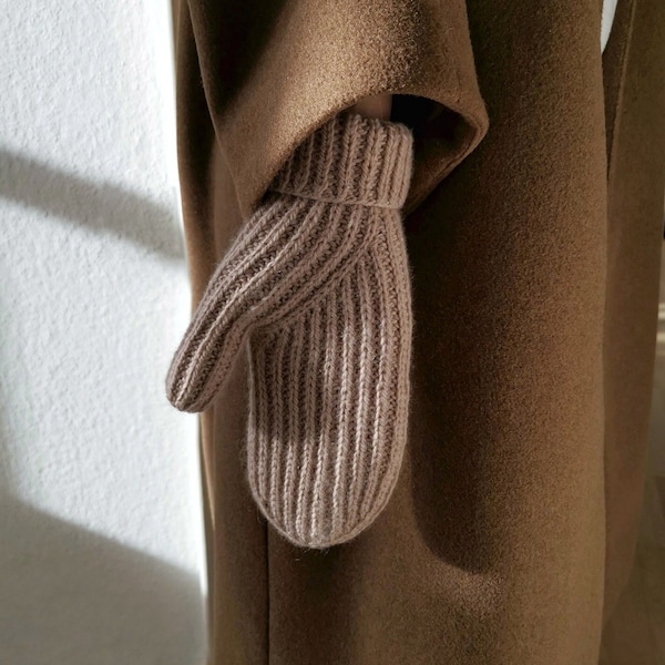 May include: A brown knitted mitten with a ribbed texture. The mitten is worn on the hand of a person wearing a brown coat.