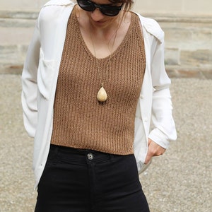 以下が含まれることがあります： 茶色のニットタンクトップ、白いボタンダウンシャツ、黒いパンツを着用した女性。彼女は、洋梨型のペンダントが付いた金のネックレスをしています。