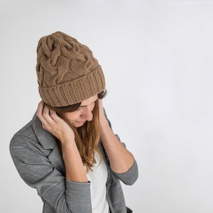 以下が含まれることがあります： 茶色のケーブルニットのビーニーとグレーの格子柄のブレザーを着た女性。彼女は下を見て、両手で頬を触っている。