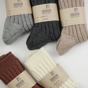 Pode incluir: Cinco pares de meias de tric&ocirc; feitas &agrave; m&atilde;o em diferentes cores, incluindo cinza, marrom, bege, branco e preto. Cada par tem uma etiqueta de papel marrom com o texto "SOCKEN HANDGESTRICKT" e espa&ccedil;o para um nome e um destinat&aacute;rio.